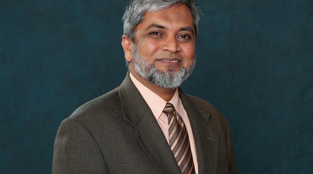 image of M. Monir Ahmed, smiling man with greying hair and facial hair, light brown skin, in brown sports coat, peach shirt and brown and silver stripped tie