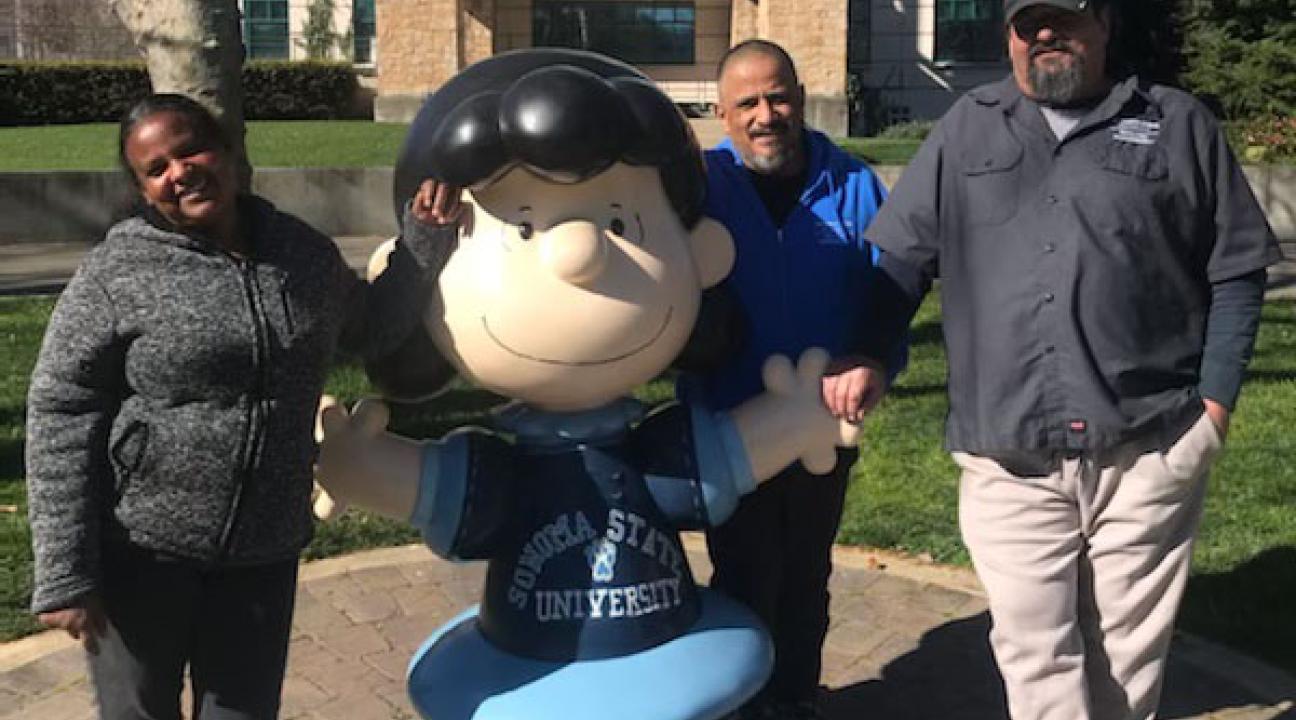 photo of three people standing next to the statue or the Peanuts character Lucy