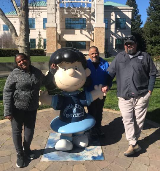 photo of three people standing next to the statue or the Peanuts character Lucy