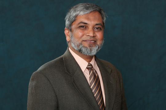 image of M. Monir Ahmed, smiling man with greying hair and facial hair, light brown skin, in brown sports coat, peach shirt and brown and silver stripped tie